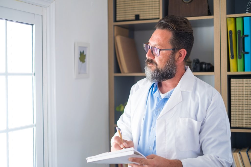 Pensive young male caucasian doctor in white medical uniform look in window